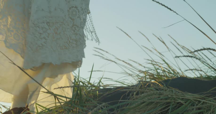 A slow-motion close-up follows a woman in a flowing white vintage dress and boots as she walks up a grassy hill at sunset