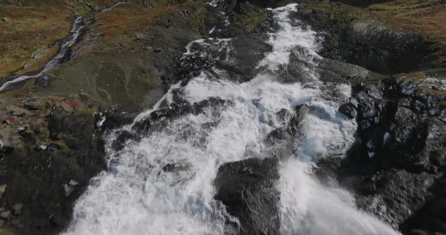 An aerial drone shot follows a powerful river as it cascades down a rugged, snowy mountainside
