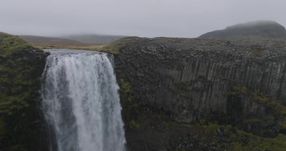 An aerial view pulls back from a powerful waterfall cascading over a dramatic basalt column cliff in a vast, overcast landscape