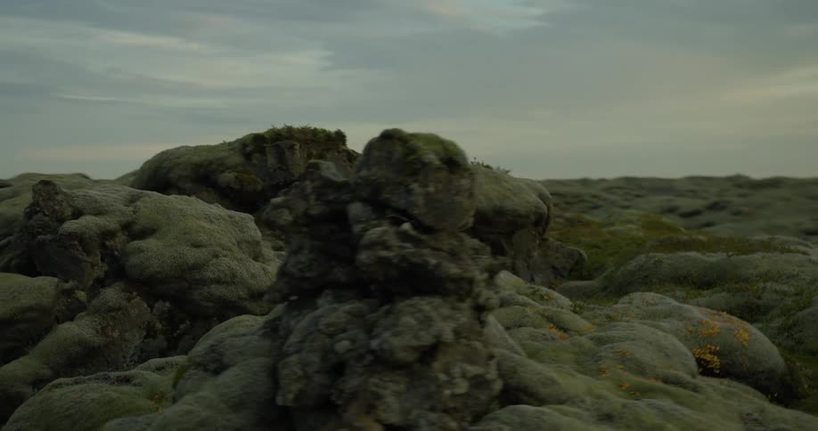 A slow, circular pan around a stone cairn amidst a vast, mosscovered volcanic landscape under an overcast sky
