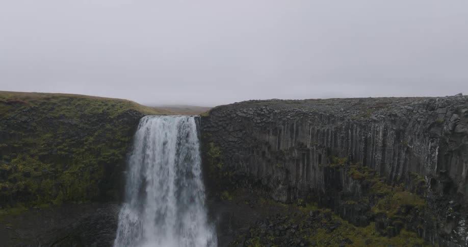A dramatic aerial shot pulls back from a powerful waterfall to reveal its source and the stunning basalt column cliffs in a misty Icelandic landscape