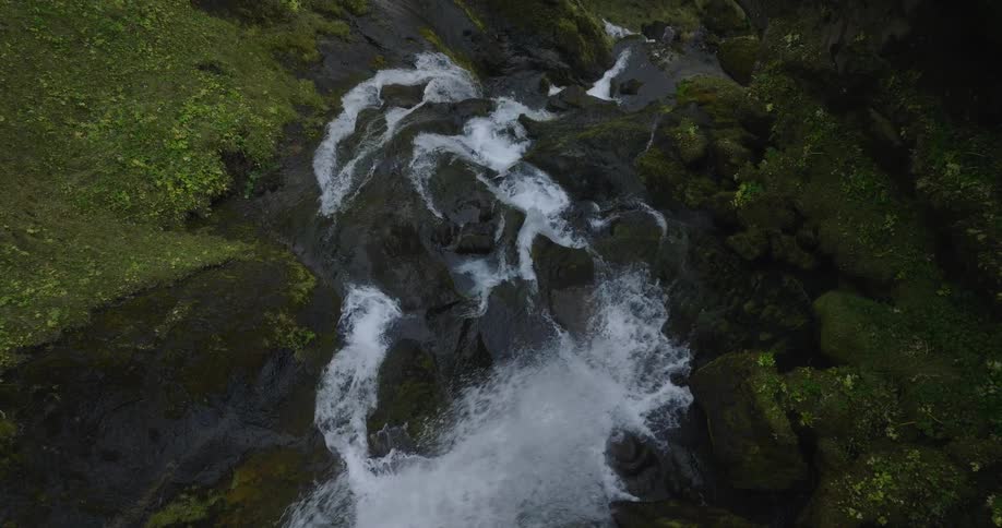 A cinematic top-down slow-motion gimbal shot reveals a beautiful waterfall cascading over dark, mossy rocks in a lush green forest
