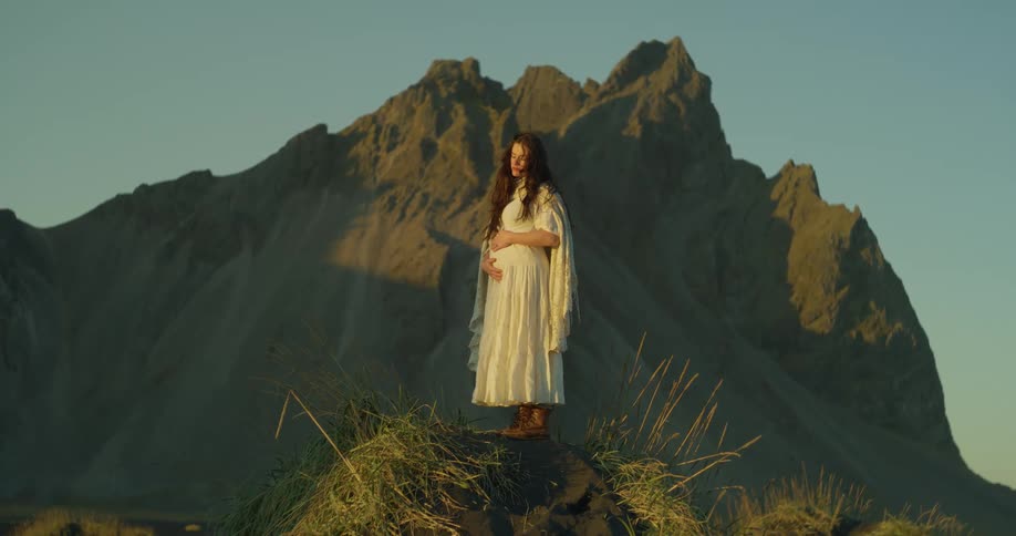 A beautiful pregnant woman in a white dress embraces her belly while standing on a hill before a majestic mountain at sunset