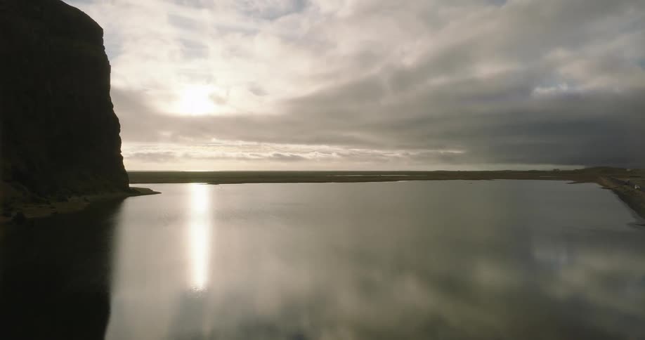 A slow aerial view pushes forward over a calm Icelandic lake, capturing the beautiful reflection of the sun breaking through moody clouds