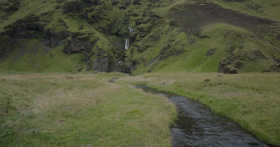 An aerial shot flies over a river through old concrete bridge ruins towards a distant waterfall cascading down a lush green Icelandic mountain