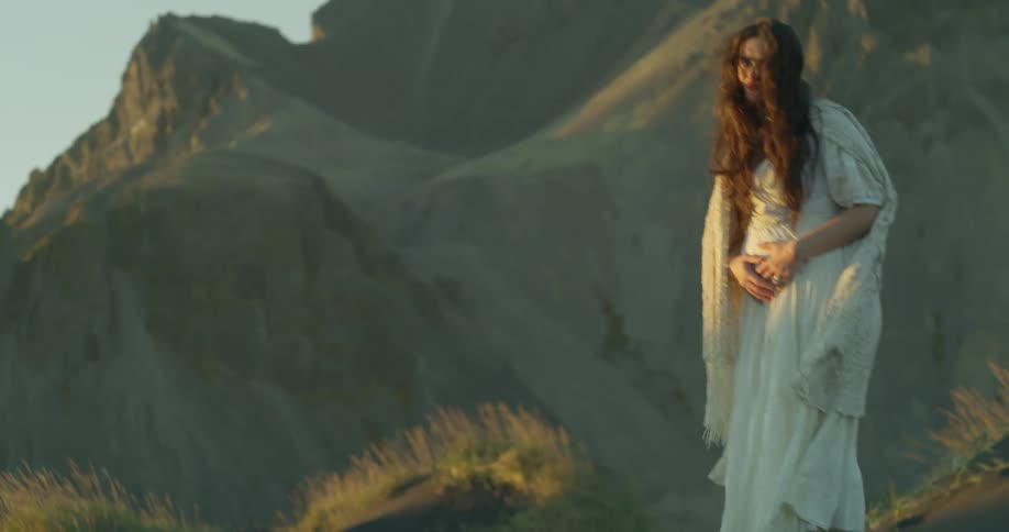 A pregnant woman in a flowing white gown peacefully embraces her belly while standing in a majestic, sunlit mountain landscape