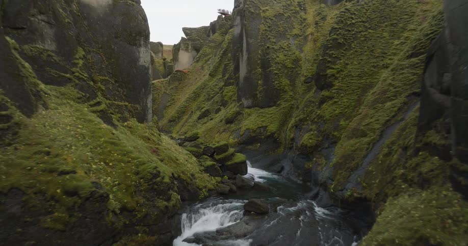 Aerial footage glides through the lush, green, and rocky Fjadrargljufur Canyon in Iceland, revealing the winding river below
