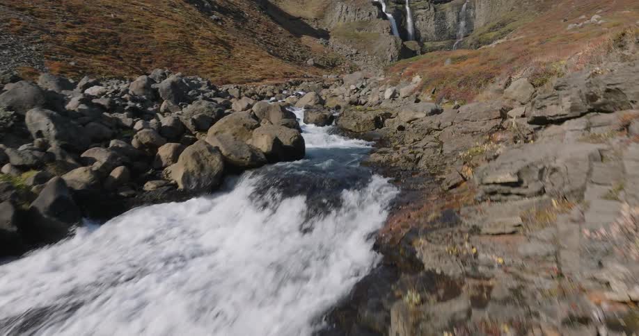 An aerial view reveals a magnificent waterfall as it glides upstream through a rocky, sunlit valley during autumn in Iceland