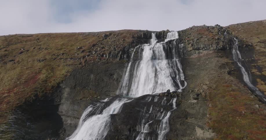 A majestic aerial shot slowly ascends a powerful, multi-tiered waterfall cascading down a rugged, rocky mountainside