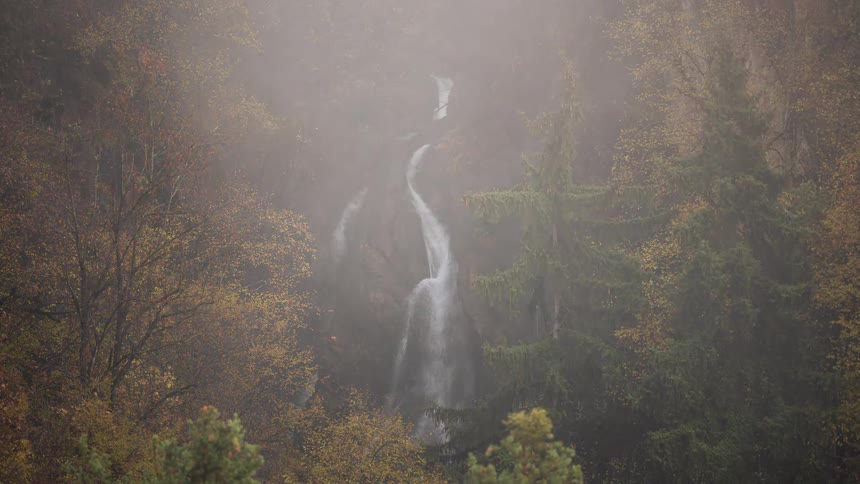 Time-lapse of dense morning fog rolling in and obscuring a beautiful waterfall in a vibrant autumn forest
