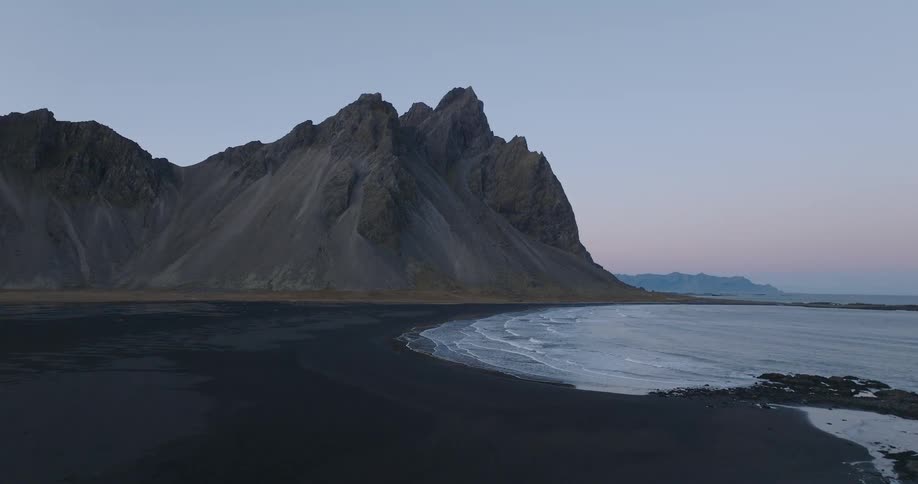 A serene, static shot of the majestic Vestrahorn mountain range meeting the black sand beach in Iceland as gentle waves wash ashore at dusk