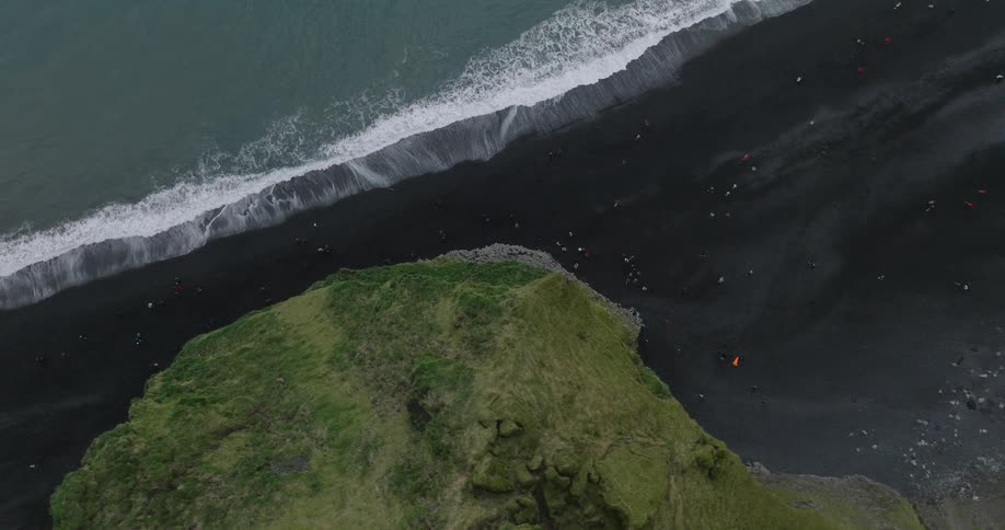 An aerial top-down view of tourists exploring the famous black sand beach next to crashing ocean waves and steep green cliffs