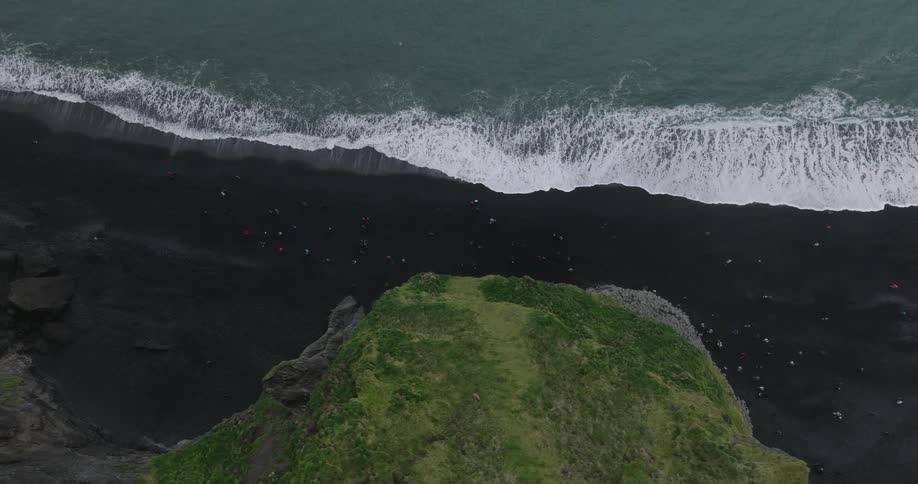 A dramatic top-down aerial shot captures tourists on a black sand beach as powerful ocean waves crash against the shore next to a verdant cliff