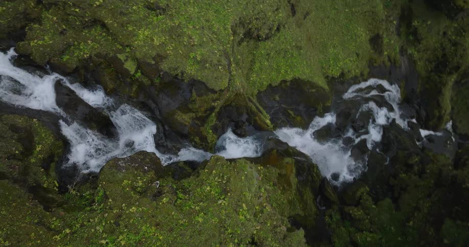 A slow-moving top-down aerial shot follows a cascading waterfall down a vibrant green, moss-covered canyon