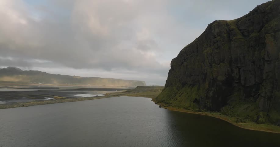 Epic aerial shot flies over a serene Icelandic lake towards a massive green cliff and a vast, dark coastal plain under a cloudy sky