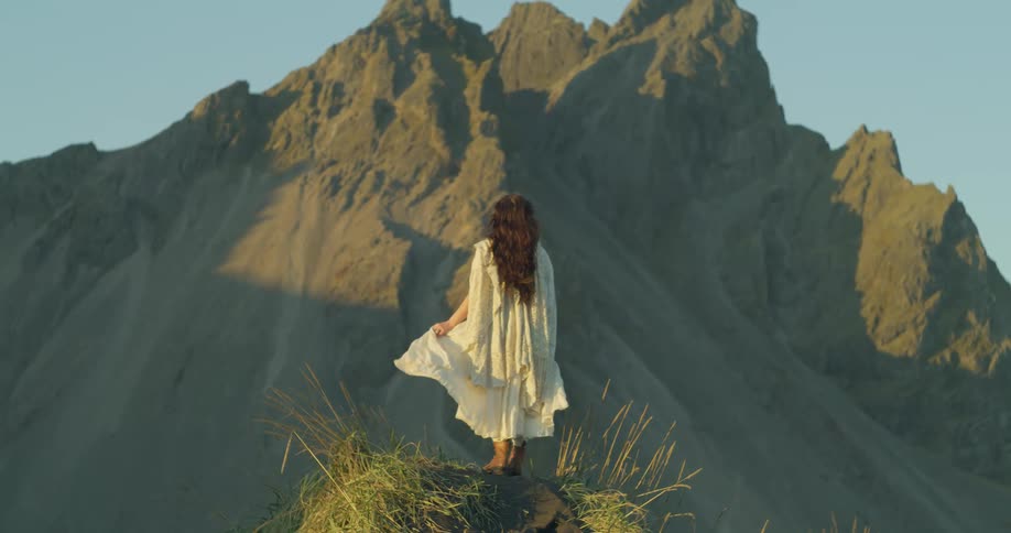 A woman in a flowing white dress stands on a grassy hill in slow motion, her hair and clothes blowing in the wind as she gazes at a majestic mountain during the golden hour