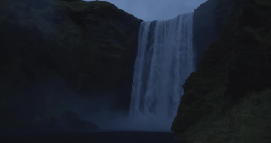 A majestic waterfall cascades down a dark, moss-covered cliff face into a misty basin at dusk