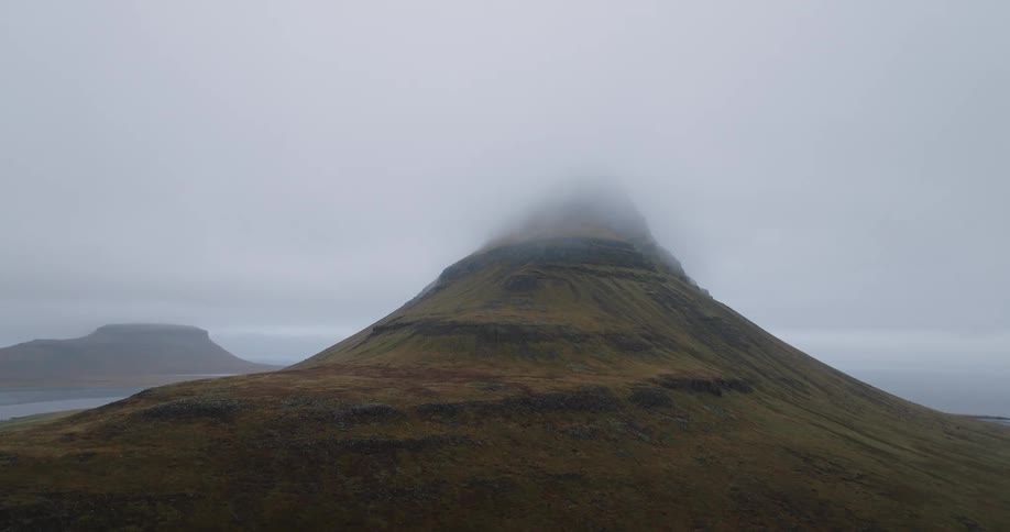 A slow aerial pullback shot reveals an iconic Icelandic mountain with its peak enveloped by dense clouds on a moody day