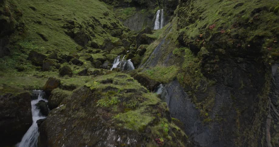A slow aerial shot follows a pristine river through a rocky, moss-covered green canyon towards a cascading waterfall
