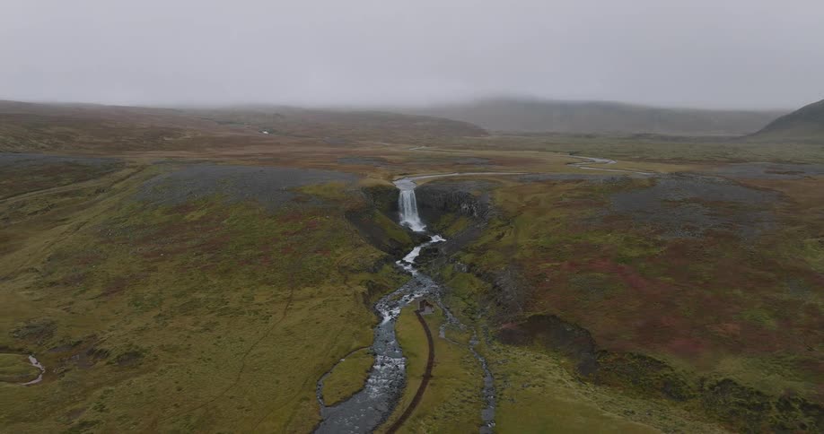 A slow aerial pullback reveals an epic waterfall cascading into a winding river through a vast, moody, and misty Icelandic valley