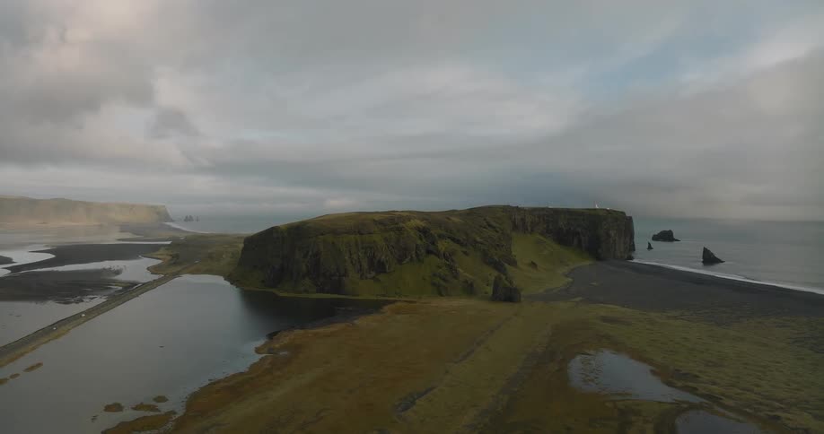 A stunning aerial view pulls back to reveal the dramatic Dyrhólaey peninsula in Iceland, surrounded by a black sand beach and the vast ocean under a cloudy sky