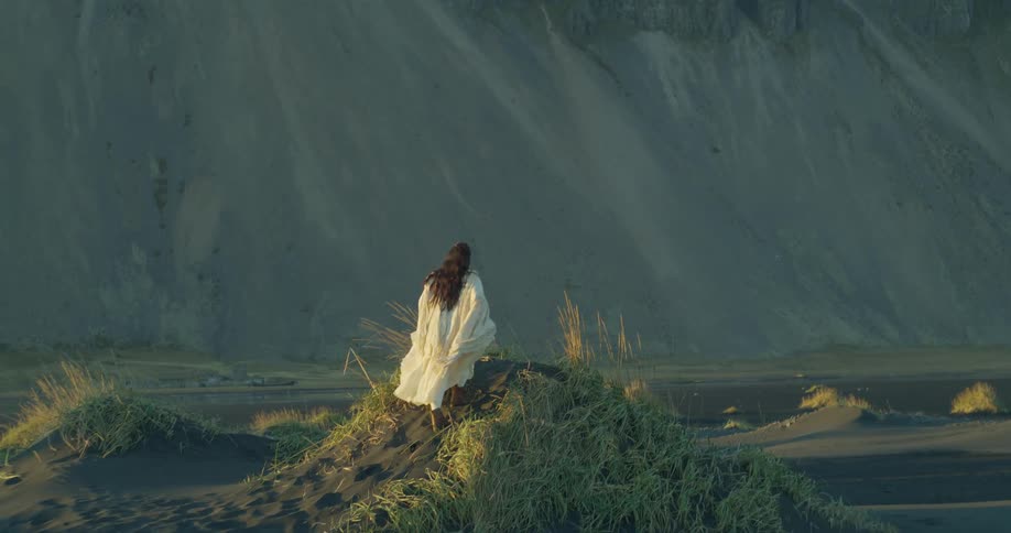 A solitary woman in a flowing white gown walks in slow motion up a grassy black sand dune, overlooking a vast mountain landscape at dusk