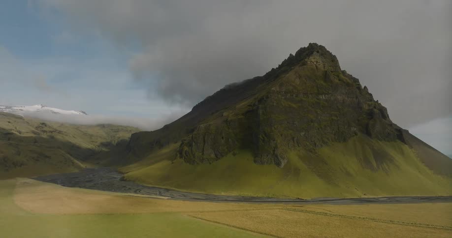 An epic aerial shot flies over green fields towards a towering, cloud-covered mountain in a vast Icelandic valley
