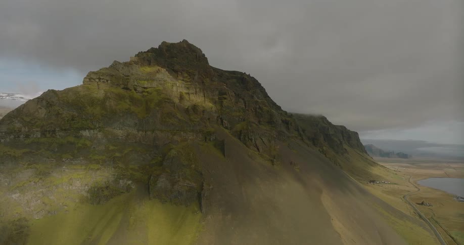 A dramatic aerial shot slowly pulls back from a sun-dappled, mossy mountain under an overcast sky, revealing a vast Icelandic landscape