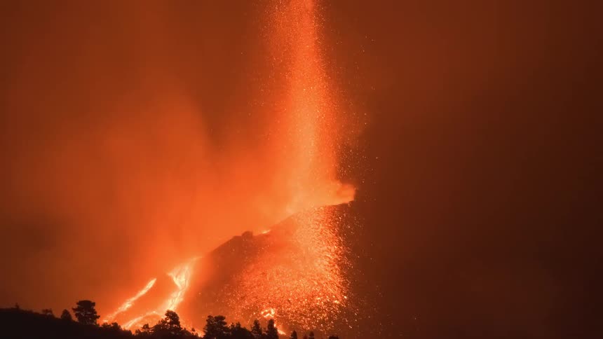 A powerful volcanic eruption at night sends a dramatic fountain of lava into the smokey, red sky