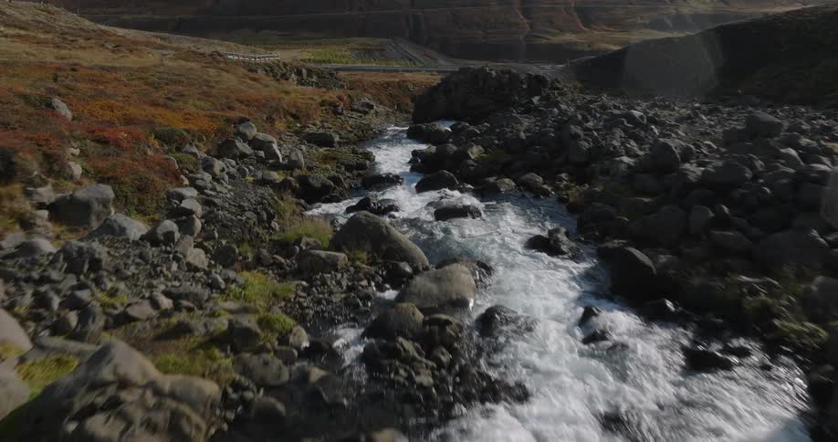 An aerial shot follows a fast-flowing rocky river upstream through a sunlit mountain valley in Iceland