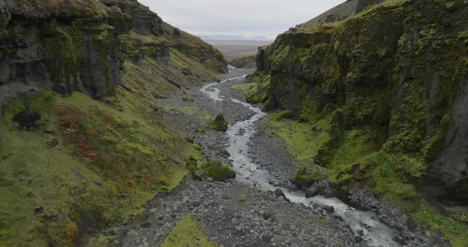 An aerial drone shot flies through a winding, moss-covered green canyon with a rocky river flowing towards the open plains of Iceland