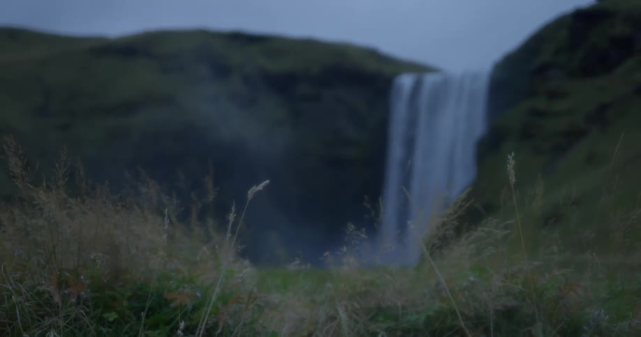 A serene shot of a majestic waterfall out of focus in the background, with tall blades of grass in focus in the foreground on an overcast day