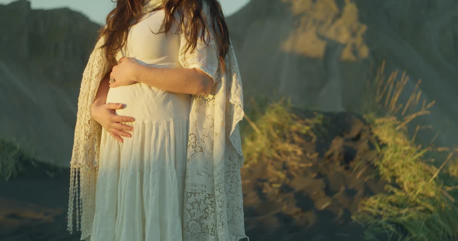 A close-up shot shows a pregnant woman in a white dress and shawl tenderly holding her baby bump in a windy, natural setting during golden hour