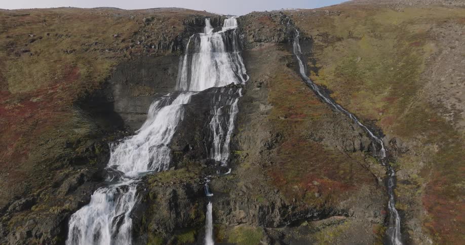 A dramatic aerial view ascends from a powerful cascade to reveal a massive, multi-tiered waterfall flowing down a rugged, mossy Icelandic mountain