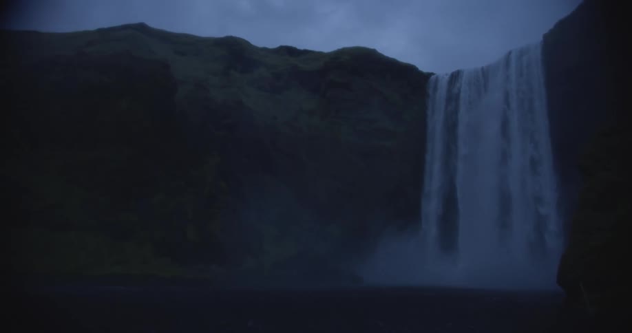 A powerful Icelandic waterfall cascades down a steep, moss-covered cliff in the deep twilight
