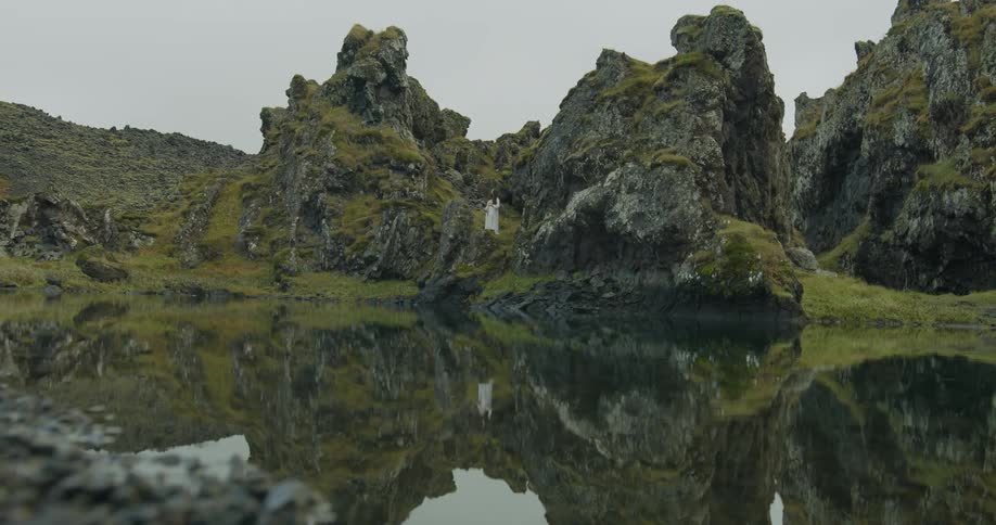 A woman in a white dress stands on a moss-covered volcanic rock formation, her reflection perfectly mirrored in the still water of a remote lake