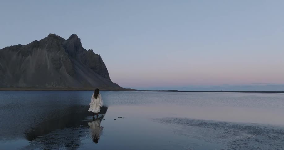 A woman in a flowing white dress walks through the calm, reflective waters of an Icelandic lagoon towards the Vestrahorn mountains at twilight