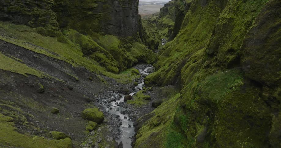 A slow aerial flight follows a winding river through a majestic, moss-covered canyon in the Icelandic highlands