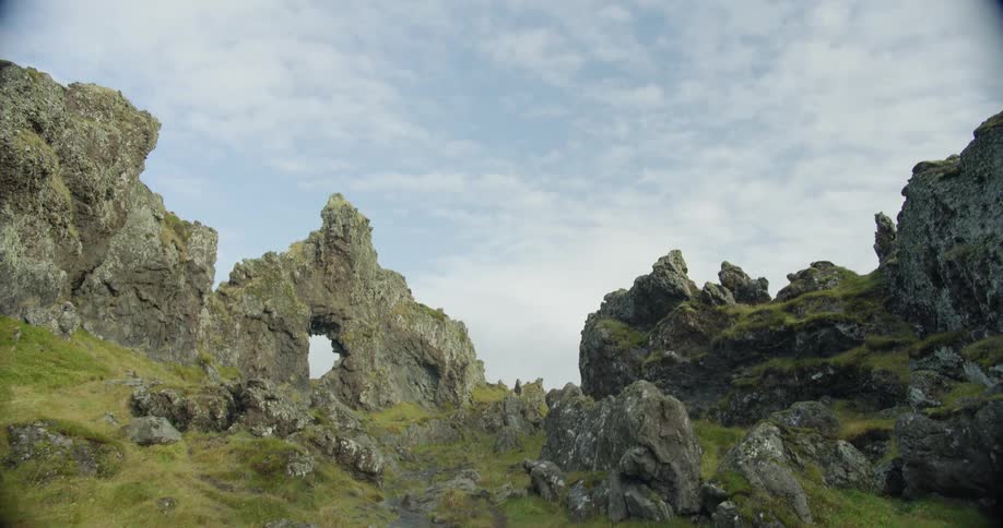 A slow tilt down from a cloudy sky reveals a majestic, rocky landscape with a natural arch