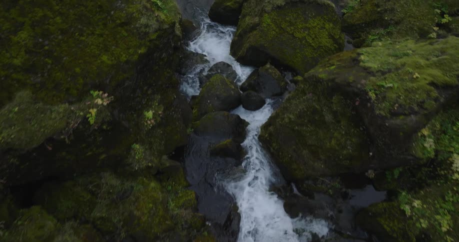 An aerial view slowly follows a stream as it cascades over dark, moss-covered rocks in a lush green valley