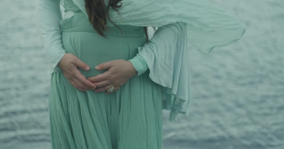 A close-up of a pregnant woman's belly with hands forming a heart shape, her flowing green dress blowing in the wind by the water