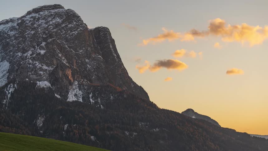 Golden hour timelapse shows clouds moving across a majestic snow-capped mountain as the sky changes from blue to a warm orange glow at sunset