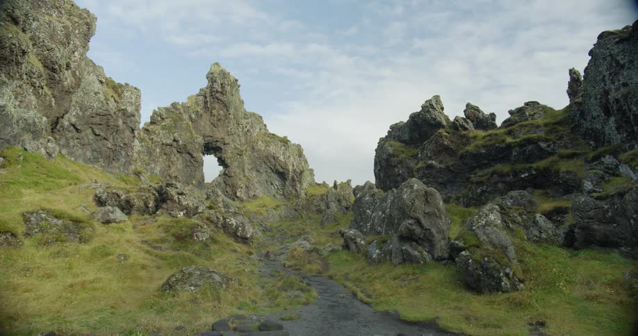 Camera pans and pulls back to reveal a rugged trail through ancient, mossy volcanic rock formations with a distinctive natural arch in Iceland