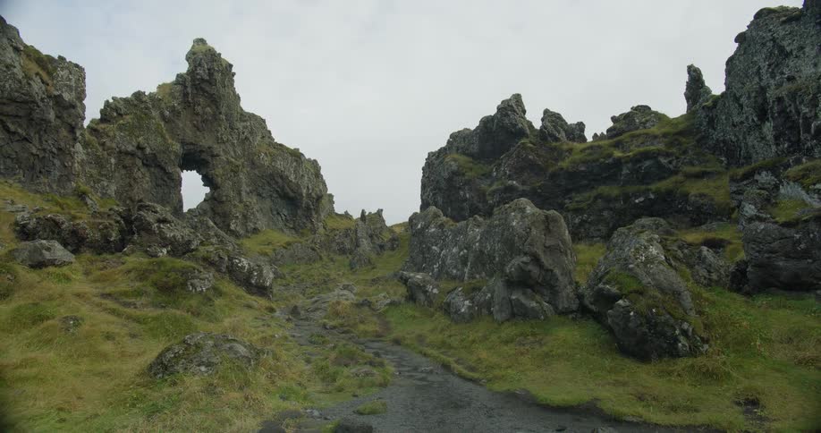 A hiking trail winds through a rugged, moss-covered volcanic rock formation with a natural arch under an overcast sky