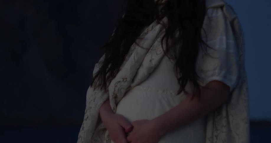 A pregnant woman in a white dress stands serenely before a dramatic mountain peak at dusk, cradling her belly
