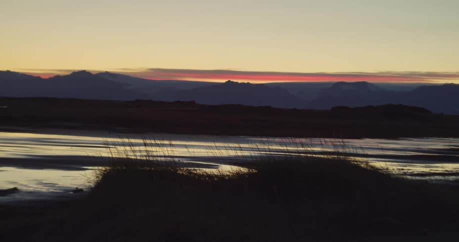 A serene view of a red sunset over distant mountain silhouettes, reflected in the calm waters of a dark, grassy marshland