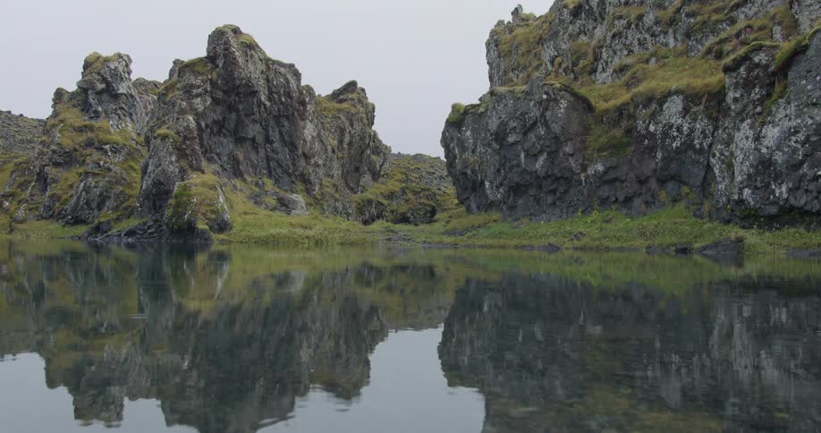 Mossy lava rock formations are perfectly reflected in a serene pond as raindrops create ripples on the still water