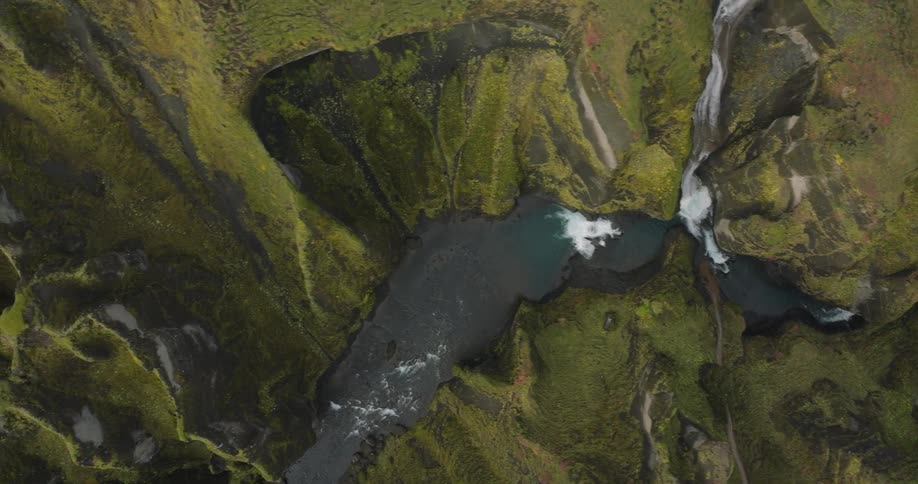 A stunning top-down aerial shot tracks a winding river through a majestic, green, moss-covered canyon with cascading waterfalls