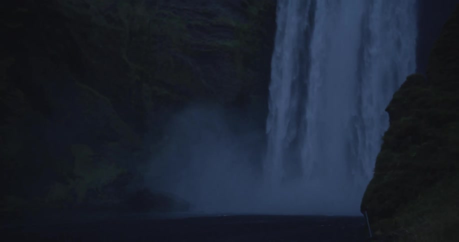 A majestic waterfall cascades down a dark, moss-covered cliff in the dim light of dusk, with the camera tilting down to the misty abyss below