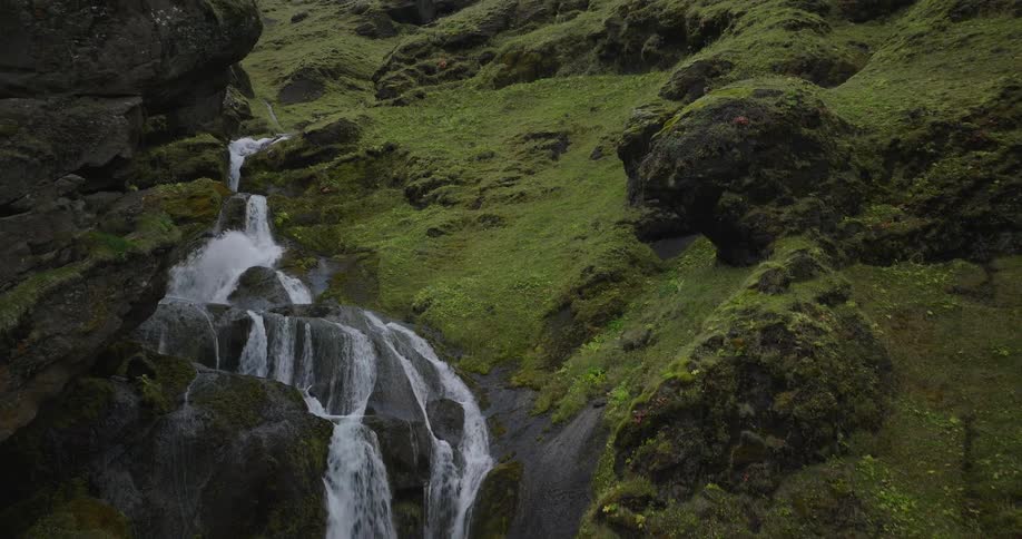 Cinematic drone footage slowly moves toward and up a beautiful waterfall cascading through a lush, green, mossy canyon in Iceland